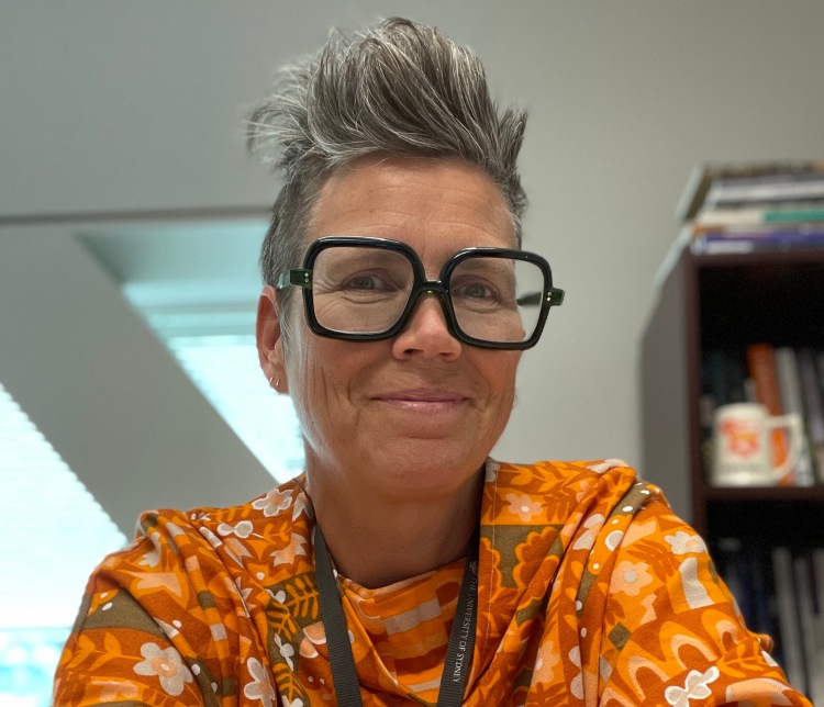 Head shot of a woman with short grey hair that is long at the top. They have bold, dark, square glasses and a small smile on their face. She is wearing an orange floral print shirt. There is the edge of a bookshelf and a triangular window in the background.