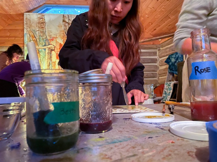 A woman with long brown hair holds a tool that makes pigment splotches on a piece of white paper. In the foreground jars of green, purple and rose pigment appear to be in use.