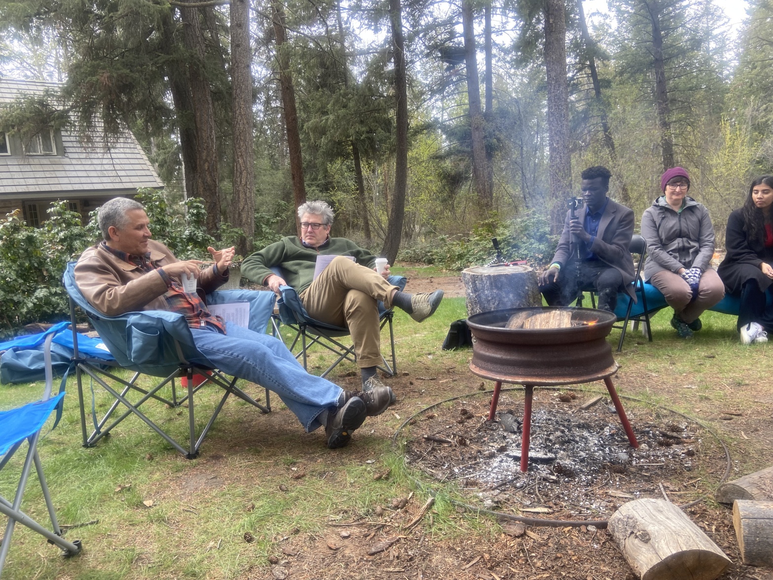 A number of individuals sit around a campfire. Two are in conversation, one films, and others listen. Trees and part of a building are in the background.