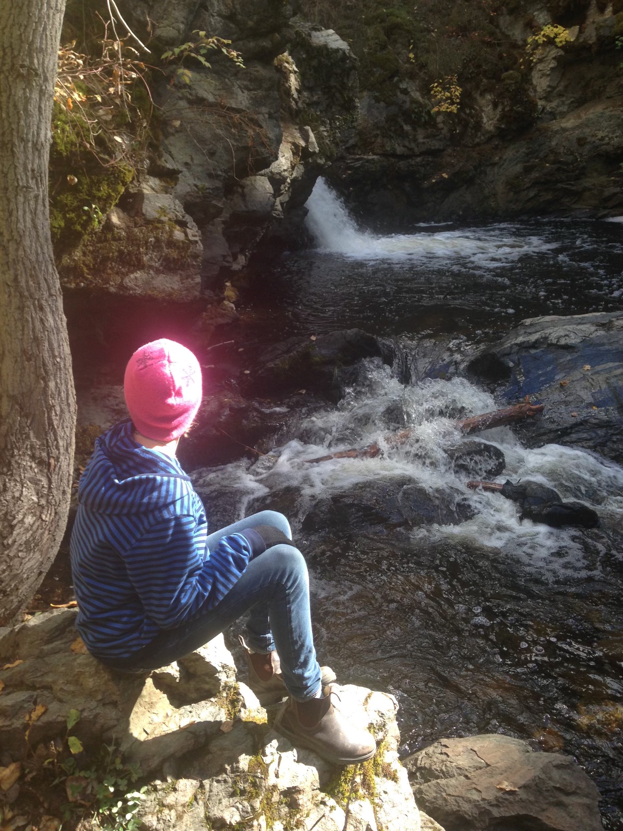 Neela sits on a rock next to the Mill Creek waterfall. The sun hits their pink hat and makes it glow.